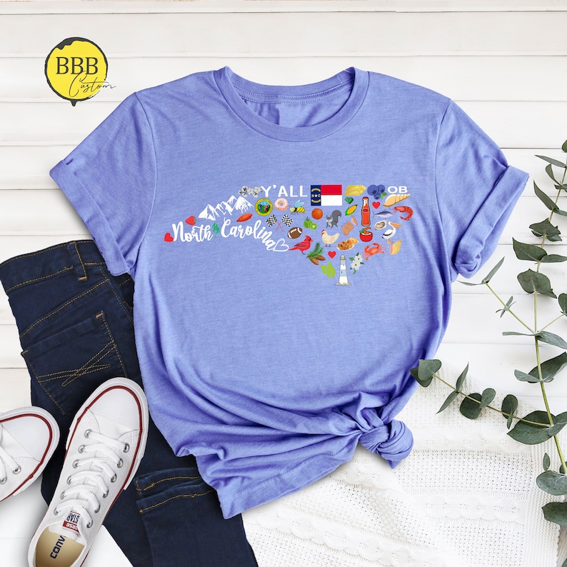 May include: A light blue t-shirt with a graphic design of a heart-shaped outline of North Carolina. The graphic includes various icons representing the state, including a lighthouse, a beach umbrella, a race car, a football, a chicken, a peach, and the state flag. The text "Y'all" and "North Carolina" are included in the graphic.