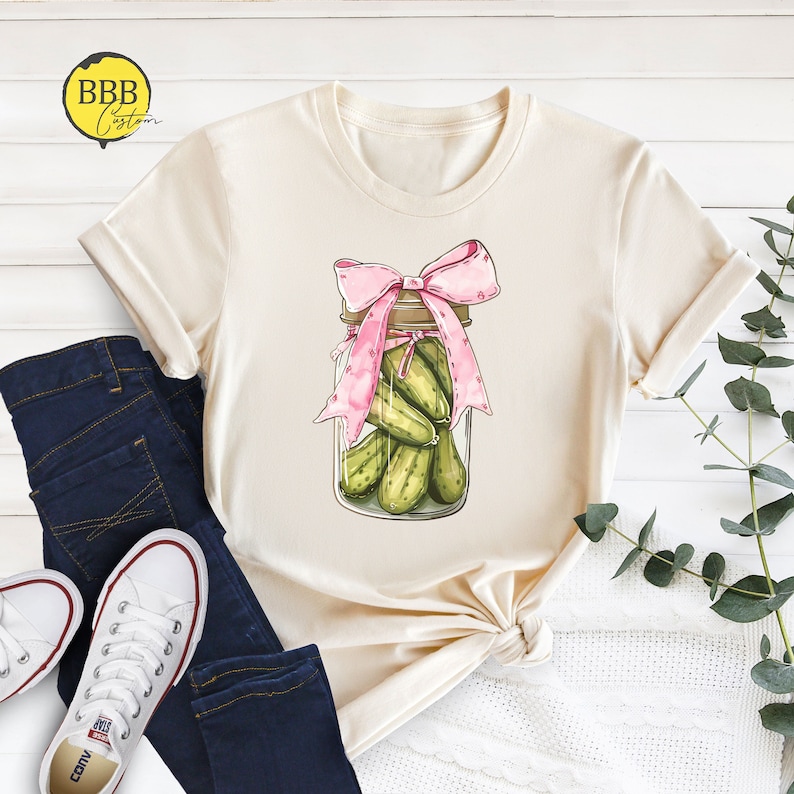 May include: A beige t-shirt with a graphic of a mason jar filled with pickles and tied with a pink ribbon.