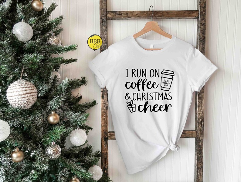 Op de afbeelding: Wit T-shirt met zwarte tekst die "I run on coffee & christmas cheer" luidt. Een koffiebeker met een sneeuwvlok is afgebeeld boven het woord "coffee". Een klein cadeaudoosje is afgebeeld naast het woord "cheer".