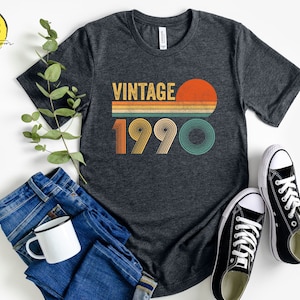 May include: A dark gray t-shirt with a vintage-style graphic that says "Vintage 1990" with a sunset and stripes in orange, yellow, green, and blue.