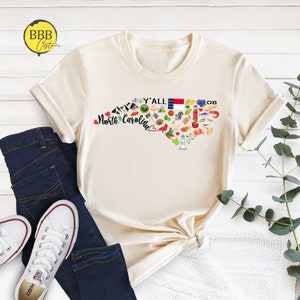 May include: A beige t-shirt with a graphic of the state of North Carolina. The graphic includes a heart shape with the words "Y'all North Carolina" and various icons representing the state, such as a lighthouse, a beach umbrella, a pig, and a basketball.