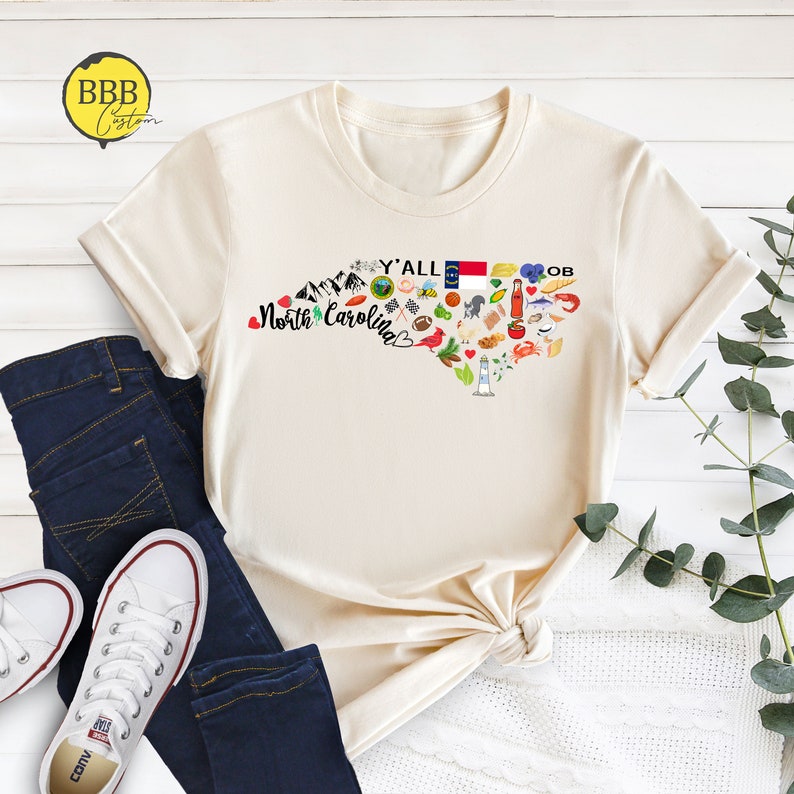 May include: A beige t-shirt with a graphic of the state of North Carolina. The graphic includes a heart shape with the words "Y'all North Carolina" and various icons representing the state, such as a lighthouse, a beach umbrella, a pig, and a basketball.