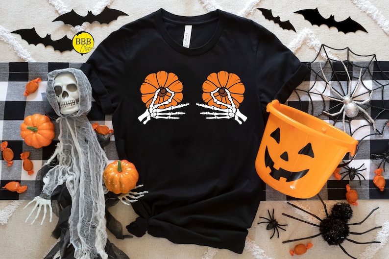 May include: Black t-shirt with a Halloween-themed design. The graphic features two orange pumpkins held by white skeleton hands. The shirt is surrounded by Halloween decorations, including pumpkins, spiders, and bats.