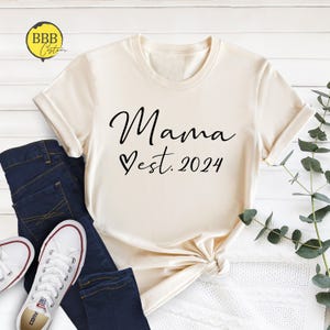 May include: A cream colored t-shirt with the text "Mama est. 2024" in black lettering with a heart symbol.