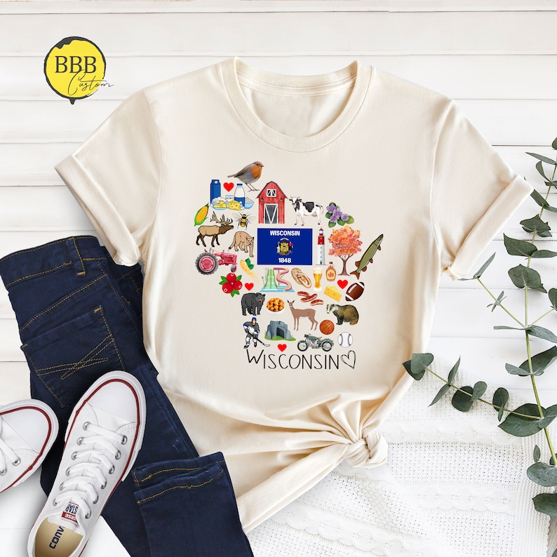 May include: A cream-colored t-shirt with a heart-shaped design featuring various icons representing Wisconsin. The icons include a red barn, a cow, a deer, a cheese wedge, a beer mug, and a football. The text "I &hearts; WISCONSIN" is printed below the heart.