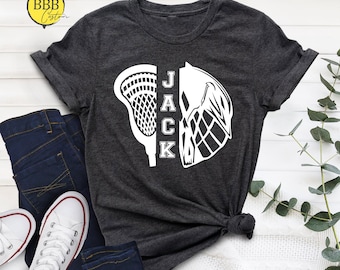 Puede incluir: Una camiseta gris oscuro con un diseño blanco de palo de lacrosse y casco. El nombre "JACK" está impreso verticalmente en la camiseta.