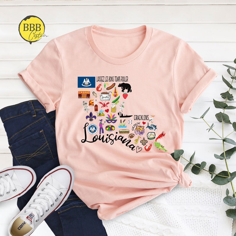 Może przedstawiać: R&oacute;żowy T-shirt z graficznym wzorem w kształcie serca przedstawiającym Luizjanę z r&oacute;żnymi ikonami reprezentującymi stan, w tym flagę stanu, lilie, raka, ciasto kr&oacute;lewskie i tekst "Laissez les bons temps rouler" i "Cracklins".