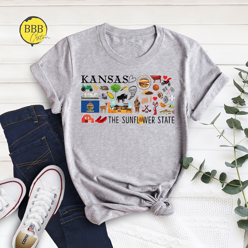 Peut inclure: Un t-shirt gris avec un motif graphique repr&eacute;sentant le mot "Kansas" et diverses ic&ocirc;nes repr&eacute;sentant l'&Eacute;tat, notamment un tournesol, un bison, une grange rouge et une tornade. Le texte "The Sunflower State" est &eacute;galement inclus sur le t-shirt.