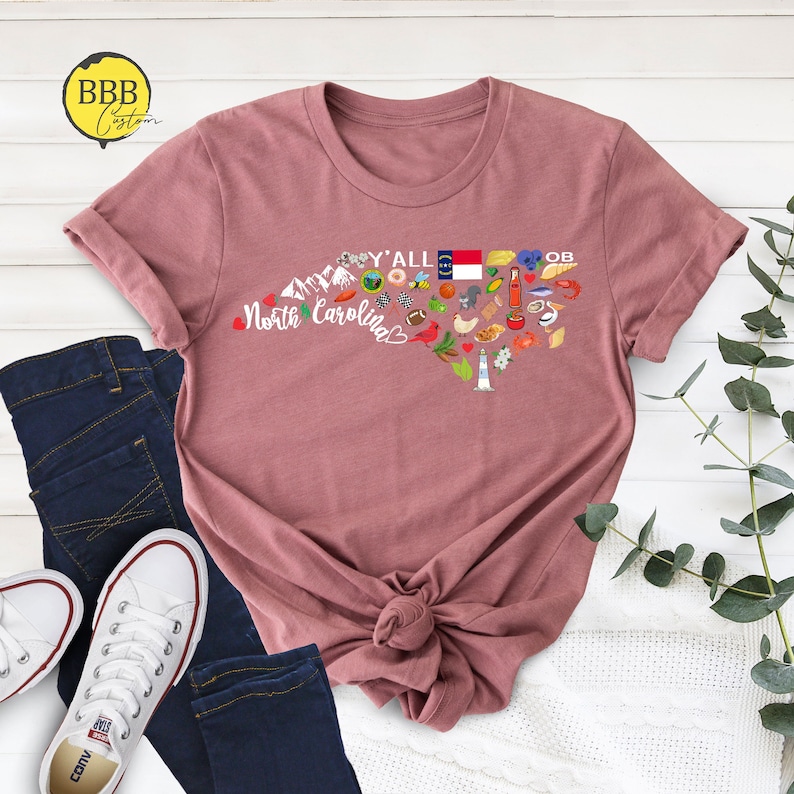 May include: A pink t-shirt with a graphic of the state of North Carolina, featuring various icons representing the state, including mountains, a lighthouse, a pig, a peach, a bee, a football, and the state flag. The text "Y'all" and "North Carolina" are also included in the graphic.