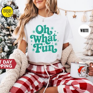 May include: A white t-shirt with the teal text "Oh What Fun", worn with red and white checked pyjama bottoms. The person holds a mug with a Santa Claus design. A decorated Christmas tree is in the background.