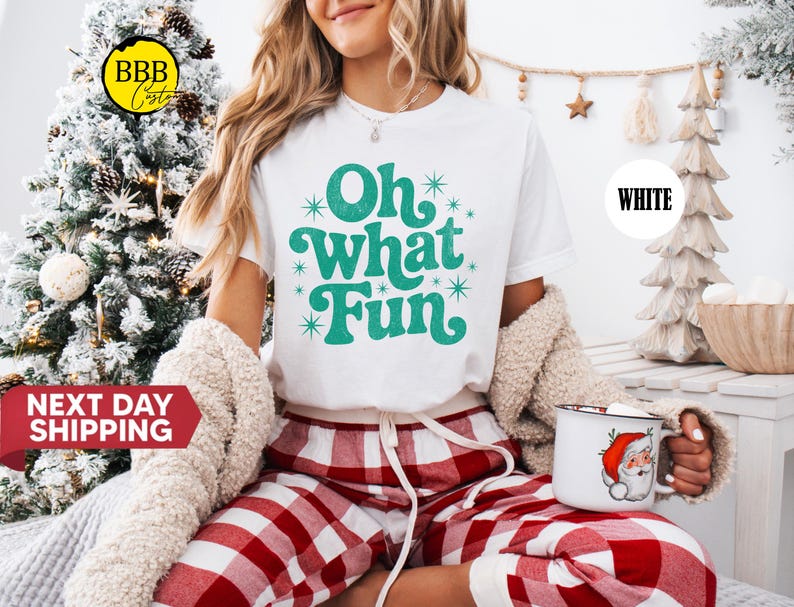 May include: A white t-shirt with the teal text "Oh What Fun", worn with red and white checked pyjama bottoms. The person holds a mug with a Santa Claus design. A decorated Christmas tree is in the background.