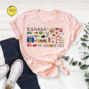Peut inclure: Un t-shirt rose avec un motif graphique noir repr&eacute;sentant le mot "Kansas" et un c&oelig;ur avec un tournesol. Le graphique comprend &eacute;galement diverses ic&ocirc;nes repr&eacute;sentant l'&Eacute;tat du Kansas, comme un champ de bl&eacute;, un bison, une tornade et une chaussure rouge. Le texte "The Sunflower State" est imprim&eacute; sous le graphique.