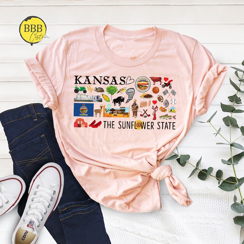 Peut inclure: Un t-shirt rose avec un motif graphique noir repr&eacute;sentant le mot "Kansas" et un c&oelig;ur avec un tournesol. Le graphique comprend &eacute;galement diverses ic&ocirc;nes repr&eacute;sentant l'&Eacute;tat du Kansas, comme un champ de bl&eacute;, un bison, une tornade et une chaussure rouge. Le texte "The Sunflower State" est imprim&eacute; sous le graphique.