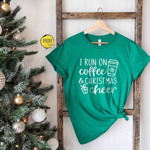 Op de afbeelding: Groen T-shirt met witte tekst die "I run on coffee & christmas cheer" luidt. Naast de tekst is een koffiebeker met een sneeuwvlok afgebeeld. Het shirt hangt aan een houten ladder voor een kerstboom.