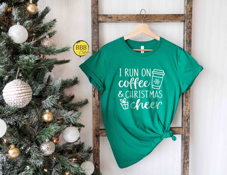 Op de afbeelding: Groen T-shirt met witte tekst die "I run on coffee & christmas cheer" luidt. Naast de tekst is een koffiebeker met een sneeuwvlok afgebeeld. Het shirt hangt aan een houten ladder voor een kerstboom.