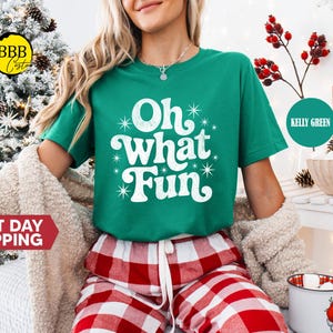 May include: A green t-shirt with the words "Oh What Fun" in white. The model is wearing red and white checked pyjama bottoms. A sign reading "North Pole Milk & Cookie" is visible in the background.