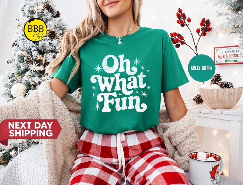 May include: A green t-shirt with the words "Oh What Fun" in white. The model is wearing red and white checked pyjama bottoms. A sign reading "North Pole Milk & Cookie" is visible in the background.