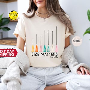 May include: Cream-colored t-shirt featuring a graphic of several syringes with colorful tips. The text "SIZE MATTERS" and the hashtag "#nurselife" are printed below the syringes. The shirt is worn by a person.