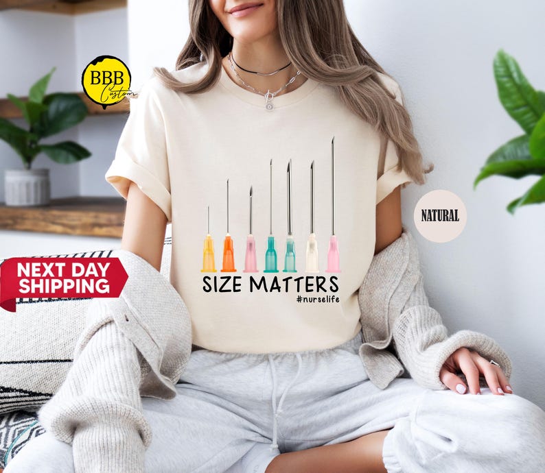 May include: Cream-colored t-shirt featuring a graphic of several syringes with colorful tips. The text "SIZE MATTERS" and the hashtag "#nurselife" are printed below the syringes. The shirt is worn by a person.