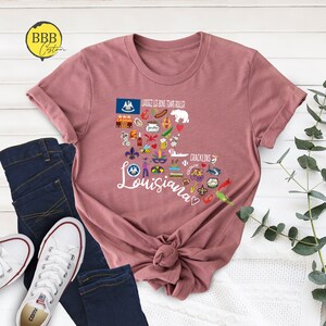 Może przedstawiać: R&oacute;żowy T-shirt z graficznym wzorem w kształcie serca przedstawiającym Luizjanę z r&oacute;żnymi ikonami i tekstem "Laissez les bons temps rouler" i "Cracklins".