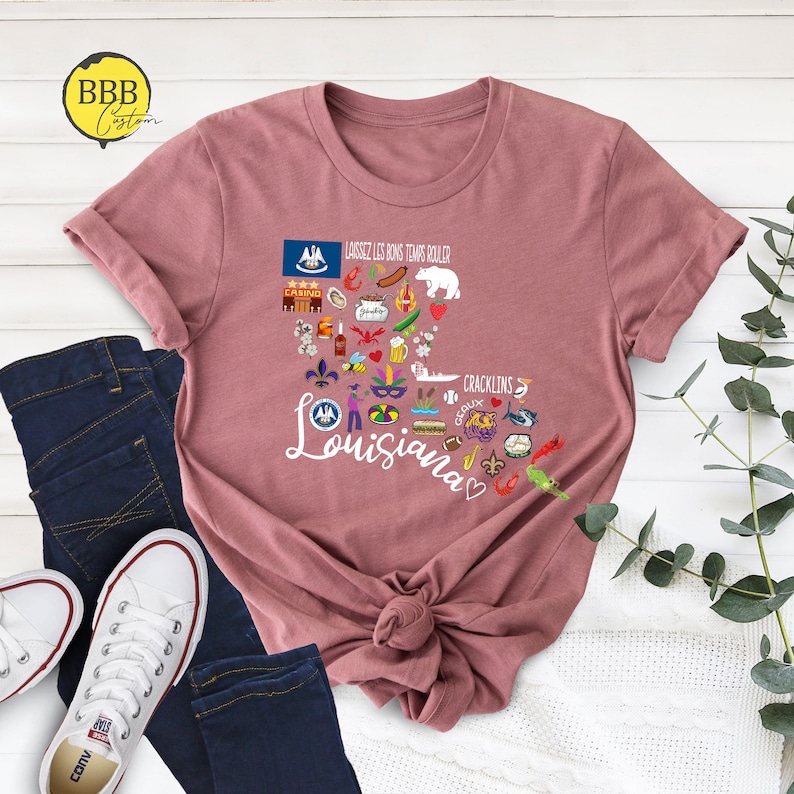 Może przedstawiać: R&oacute;żowy T-shirt z graficznym wzorem w kształcie serca przedstawiającym Luizjanę z r&oacute;żnymi ikonami i tekstem "Laissez les bons temps rouler" i "Cracklins".