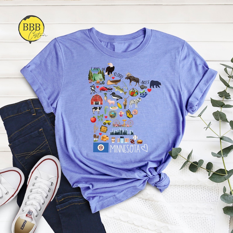 以下が含まれることがあります： ミネソタ州のグラフィックが描かれた薄いブルーのTシャツ。グラフィックには、ムース、湖、キャビン、メープルリーフなど、州の文化を表すさまざまなアイコンが含まれています。グラフィックには、「Land of 10,000 Lakes」と「Minnesota」のテキストも含まれています。