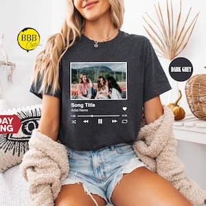 May include: Dark grey t-shirt featuring a framed image with three people and a music player interface. The text on the shirt reads "Song Title" and "Artist Name". The model is wearing denim shorts and a cream-colored sweater.