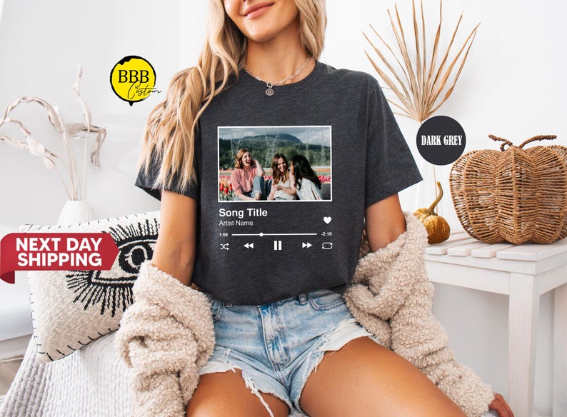 May include: Dark grey t-shirt featuring a framed image with three people and a music player interface. The text on the shirt reads "Song Title" and "Artist Name". The model is wearing denim shorts and a cream-colored sweater.