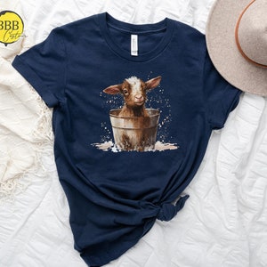 May include: A navy blue t-shirt with a graphic of a brown and white baby goat sitting in a metal bucket. The goat is surrounded by white splatters, suggesting water.