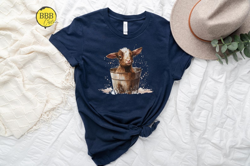 May include: A navy blue t-shirt with a graphic of a brown and white baby goat sitting in a metal bucket. The goat is surrounded by white splatters, suggesting water.