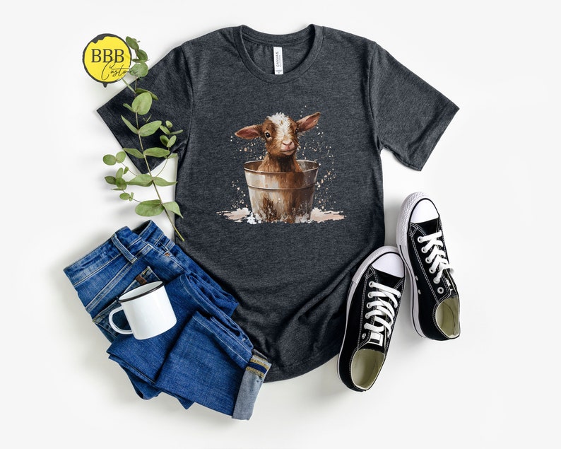 May include: A dark gray t-shirt with a graphic of a brown baby goat sitting in a bucket. The goat is looking at the camera and has a white spot on its forehead. The bucket is brown and has a white rim. The t-shirt is on a white background.
