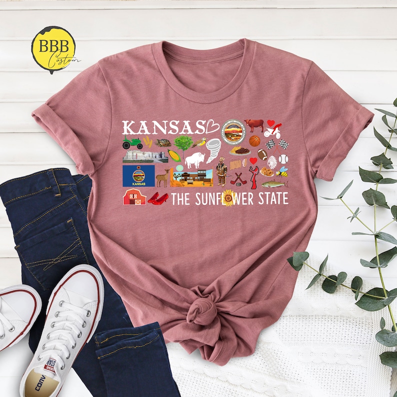 Peut inclure: Un t-shirt rose avec un motif graphique blanc repr&eacute;sentant le mot "Kansas" et un symbole de c&oelig;ur. Le graphique comprend &eacute;galement diverses ic&ocirc;nes repr&eacute;sentant des choses associ&eacute;es &agrave; l'&Eacute;tat du Kansas, comme un tournesol, un champ de bl&eacute;, un chapeau de cowboy et une pantoufle rouge en rubis. Le texte "The Sunflower State" est &eacute;galement inclus dans le graphique.