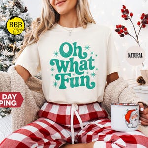 May include: A cream-coloured t-shirt with the phrase "Oh What Fun" in teal script and starburst accents. The person is wearing red and white plaid pyjama bottoms and holding a Christmas-themed mug with a Santa Claus design.