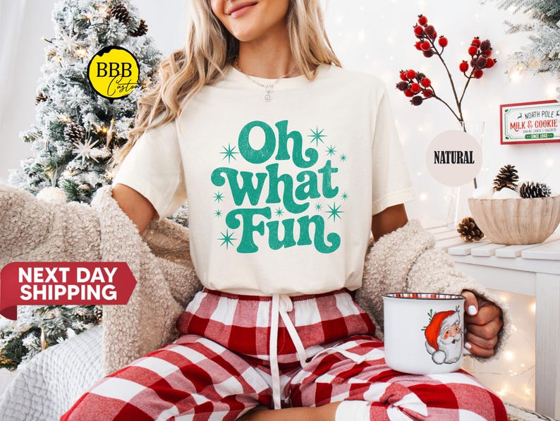 May include: A cream-coloured t-shirt with the phrase "Oh What Fun" in teal script and starburst accents. The person is wearing red and white plaid pyjama bottoms and holding a Christmas-themed mug with a Santa Claus design.