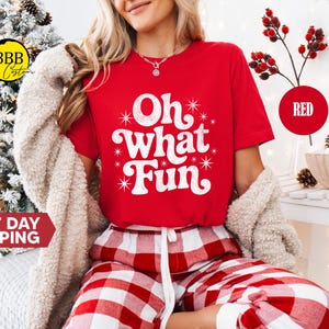 May include: A red t-shirt with the phrase "Oh what Fun" in white script. The model is wearing red and white plaid pajama bottoms and a cream-coloured cardigan. The image has a Christmas theme.