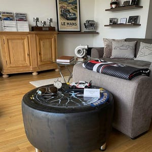 Könnte beinhalten: Ein runder Couchtisch aus einem schwarzen Michelin-Rennreifen mit Glasplatte und silbernem Felgendesign. Auf dem Tisch liegen eine Fliegerbrille, eine silberne Stoppuhr, ein weißes Modellrennauto und ein kleines weißes Schild mit Text.