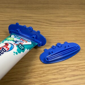 May include: A blue plastic toothpaste tube squeezer with a tube of Odol-med3 toothpaste. The toothpaste tube is white with blue and red accents and the text "6-13 Jahre". The squeezer is on a wooden surface.