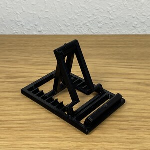 Könnte beinhalten: Ein schwarzer, 3D-gedruckter Telefonständer. Er hat eine rechteckige Basis mit Schlitzen und eine abgewinkelte Halterung zum Halten eines Telefons. Die Halterung ist schwenkbar. Hergestellt aus Kunststoff.