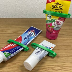 May include: Three tubes of product, including Colgate toothpaste, a yogurt dessert, and Sensodyne toothpaste, are held by green plastic tube squeezers. The products are on a wooden surface.