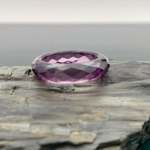 Può includere: Un anello sfaccettato color ametista con un design geometrico. L'anello è appoggiato su un pezzo di legno stagionato, che mostra la sua tonalità viola intenso e il taglio intricato. Il design dell'anello cattura la luce.