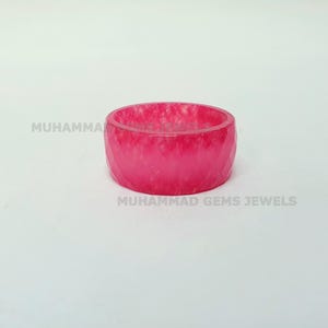 May include: A vibrant pink ring with a faceted design. The ring is a solid band of translucent material, showcasing a marbled effect. The text "MUHAMMAD GEMS JEWELS" is visible.