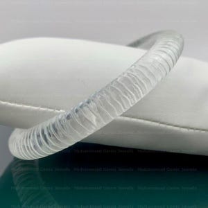 Könnte beinhalten: Ein klarer, strukturierter Armreif. Das Armband hat ein geriffeltes Design und besteht aus einem transparenten Material. Der Armreif liegt auf einer weißen Oberfläche. Das Gesamtthema ist Schmuck.