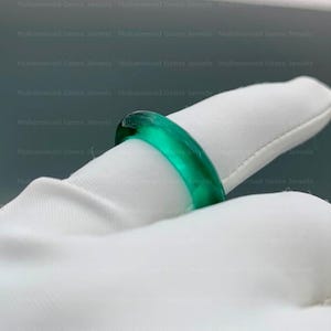 May include: A vivid, emerald-green ring. The ring is a solid band of translucent green material, with a smooth, polished surface. The ring is displayed on a white surface, accentuating its colour and design. The ring is a piece of jewellery.