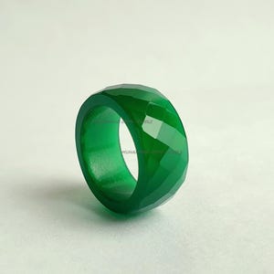 Może przedstawiać: Fasetowany, szmaragdowozielony pierścionek. Pierścionek ma szeroką obrączkę i geometryczny wzór. Pierścionek wykonany jest z półprzezroczystego materiału i umieszczony na białym tle. Biżuteria.