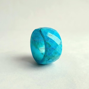 Puede incluir: Un anillo facetado de color turquesa. El anillo tiene una banda de diferentes tonos de azul y turquesa. El anillo está hecho de un material translúcido y las facetas crean un efecto brillante. Una pieza audaz.