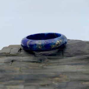 Puede incluir: Un anillo azul oscuro con motas doradas. El anillo está hecho de un material liso y pulido y se asienta sobre una superficie oscura y texturizada. El color del anillo es un azul profundo y rico con un brillo sutil.