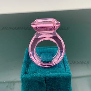 May include: A transparent pink ring with a large, rectangular gemstone. The ring is set on a teal velvet surface. The text "MUHAMMAD JEWELS" is visible in the background.