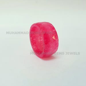 Op de afbeelding: Een helder roze ring met een facetontwerp. De ring is een effen band met een geometrisch patroon. De tekst "MUHAMMAD GEMS JEWELS" staat op de ring. De ring staat tegen een witte achtergrond.