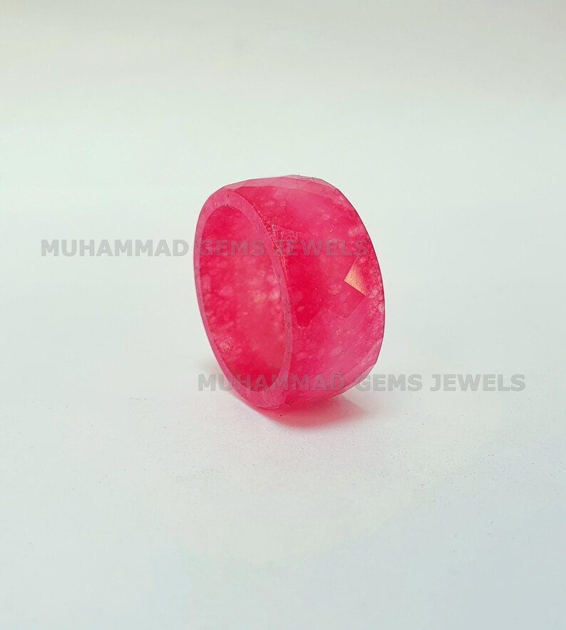 May include: A faceted, bright pink ring. The ring is a solid band with a geometric design. The text "MUHAMMAD GEMS JEWELS" is printed on the ring. The ring is set against a white background.