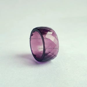 May include: A faceted, amethyst-colored gemstone ring. The ring is a deep purple hue with a geometric cut, reflecting light. The ring's design is a thick band, showcasing the gemstone's color and clarity. The ring is a statement piece.